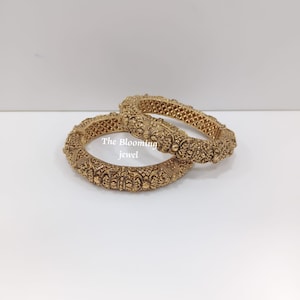 Peut inclure: Deux bracelets rigides en métal doré ornés de motifs floraux et géométriques en relief sur toute la surface. Le texte "The Blooming jewel" est imprimé en blanc au centre des bracelets, qui présentent une forme circulaire et détaillée.