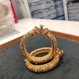 May include: Two gold-toned bangles featuring intricate embossed floral and geometric patterns. Each bangle has a prominent decorative clasp mechanism at the top with a small central flower detail, showcasing a textured metallic finish throughout.