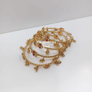 Può includere: Un set di bracciali rigidi in metallo dorato con un design a corda intrecciata, impreziositi da piccoli motivi floreali con pietre rosse e trasparenti e ciondoli pendenti. Il testo "The Blooming Jewel" è stampato al centro dei bracciali.