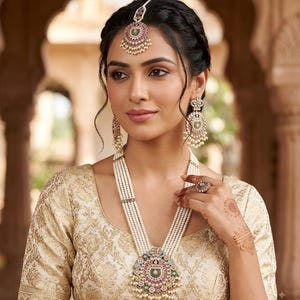 May include: A multi-strand pearl necklace with a large circular pendant featuring pink, green, and white stones, matching drop earrings, and a maang tikka headpiece. The jewelry set is worn with a cream-colored blouse with a gold brocade pattern.