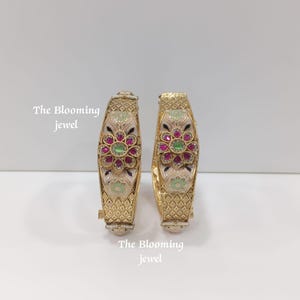 May include: Two gold-toned bangles featuring a central floral motif with pink and green stones, surrounded by cream-coloured enamel detailing and a geometric diamond-patterned texture. The text The Blooming jewel is visible on the image.