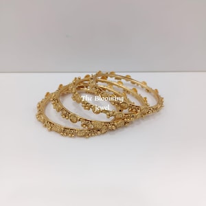 May include: A set of three gold-toned bangles featuring a textured surface with small circular accents and dangling beads. The text The Blooming Jewel is visible in the centre of the bangles. Each bangle has a consistent metallic finish.