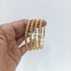 May include: A set of four gold-toned bangles featuring a repeating marquise-shaped pattern with embedded clear stones. Each bangle has a textured metal border. The text The Blooming jewel is visible across the centre of the bangles.