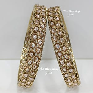 Può includere: Due bracciali rigidi color oro con un motivo ripetuto di pietre trasparenti a forma di goccia incastonate in una cornice metallica. Il testo The Blooming jewel è visibile nell'immagine. I bracciali hanno un design traforato e strutturato.