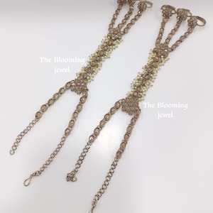 May include: Two ornate hand harnesses featuring metallic gold-tone settings with embedded teardrop-shaped crystals and small white beads. Each piece includes three ring loops connected to a central floral-shaped ornament and a wrist chain. Text reads The Blooming jewel.