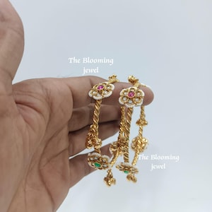 Peut inclure: Deux bracelets joncs dorés avec un design de bande métallique torsadée. Chaque bracelet est orné d'accents en forme de fleur contenant de petites perles blanches et une pierre centrale rose ou verte. Le texte The Blooming jewel est visible.