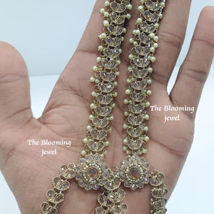 May include: A pair of long, gold-toned metal earrings featuring a floral stud design at the top, connected to a vertical chain of teardrop-shaped crystals and small white beads. The text The Blooming jewel is visible on the image.