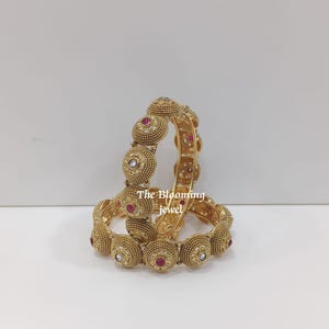 Op de afbeelding: Twee goudkleurige armbanden met een getextureerd, korrelig oppervlak en cirkelvormige motieven. Elk cirkelvormig element is geaccentueerd met een kleine, centrale rode of witte steen. De tekst The Blooming jewel is zichtbaar in het midden.