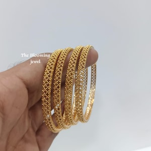 May include: A set of four gold-toned bangles featuring a detailed lattice or mesh pattern across the surface. The text The Blooming jewel is visible in the upper left corner. Each bangle has a consistent circular shape and a metallic finish.