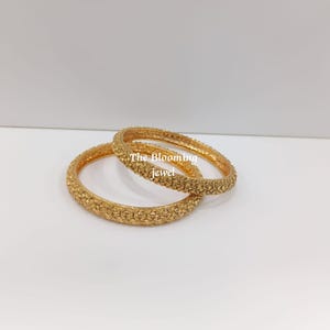 May include: Two gold-toned bangles featuring a textured, intricate floral pattern across the entire surface. The text "The Blooming jewel" is printed in white letters across the centre of the bangles, which are positioned overlapping each other.