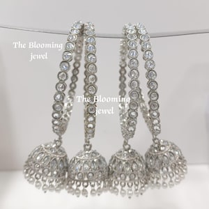 May include: Four silver-toned bangles with circular clear stones and bell-shaped jhumka pendants featuring intricate stone detailing and small dangling beads. The text The Blooming Jewel is visible on the image. These circular ornaments are metallic.