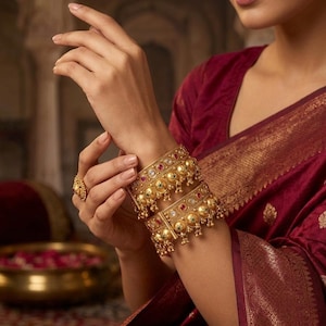 Pode incluir: Dois braceletes largos de ouro com uma série de contas esféricas em relevo e pedras incrustadas alternadas em vermelho e verde. Os braceletes são adornados com pequenos pingentes dourados pendurados na borda inferior, com um anel de ouro a combinar.