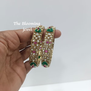 Op de afbeelding: Twee goudkleurige armbanden met een herhalend bloemmotief, ingelegd met heldere stenen, ovale roze stenen en driehoekige groene accenten. De tekst The Blooming jewel is zichtbaar in de linkerbovenhoek van de afbeelding.