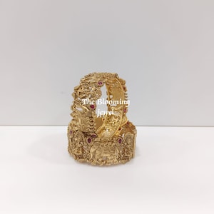 Può includere: Due bracciali rigidi color oro con intricati motivi in rilievo raffiguranti elefanti e divinità, arricchiti da piccole pietre rosse a goccia incastonate. Il testo The Blooming Jewel è sovrapposto in bianco al centro dei bracciali.