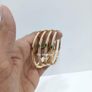 Può includere: Un set di quattro bracciali rigidi color oro con una fila centrale di piccole perline bianche e pietre ovali alternate rosse e verdi. Il testo The Blooming Jewel è visibile sui bracciali. Il design presenta un bordo con una texture.