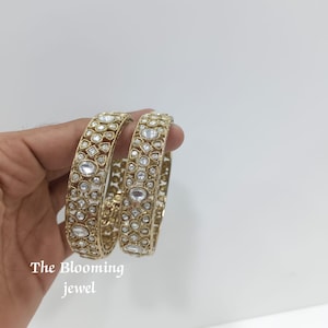 May include: Two gold-toned bangles featuring an intricate filigree pattern embedded with numerous clear, faceted stones in various shapes. The text 'The Blooming jewel' is printed in white in the lower left corner of the image.