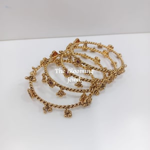 May include: A set of gold-toned metal bangles featuring a twisted rope design. Each bangle is adorned with small, dangling charms and embedded with tiny red stones. The text "The Blooming jewel" is overlaid in the centre of the stacked bangles.