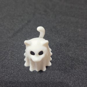 Peut inclure: Petite figurine blanche en forme de chat fantôme avec des yeux noirs et une queue recourbée. Le fantôme a un corps drapé et froissé. La figurine est posée sur un fond gris foncé.