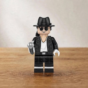 May include: A plastic minifigure wearing a black fedora, black sunglasses, and a black jacket with white speckles over a white shirt. The figure has black hair, white gloves, and black pants with white accents, and is holding a silver microphone.