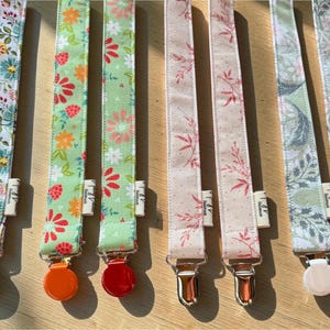 May include: Eight fabric pacifier clips arranged in pairs, featuring various floral and botanical patterns in shades of blue, red, green, pink, and grey. Each strap includes a metal or plastic clip at the bottom and a small white fabric tag.
