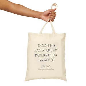 May include: A beige canvas tote bag with two top handles. The front features black text that reads: DOES THIS BAG MAKE MY PAPERS LOOK GRADED? Below this, the text reads: Mrs. Smith Washington Elementary in a cursive script font.