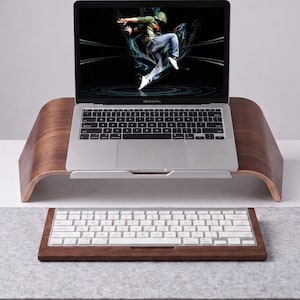 May include: A curved wooden laptop stand holding a silver laptop with a black keyboard and a screen displaying a person dancing. Below the stand sits a wireless keyboard with white keys and a dark wood frame, all resting on a grey felt mat.