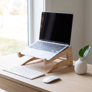 Puede incluir: Un soporte para portátil de madera clara que sostiene un portátil plateado en posición abierta. El soporte presenta un diseño curvo de dos piezas entrelazadas. Un teclado inalámbrico blanco y un ratón de ordenador blanco están colocados delante.
