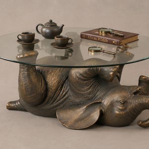 Könnte beinhalten: Ein runder Couchtisch mit Glasplatte, der von einer bronzefarbenen Skulptur eines liegenden Elefanten getragen wird. Auf der Tischplatte stehen eine dunkle Teekanne, zwei passende Tassen, ein Buch mit dem Titel African Wildlife und zwei Lupen.