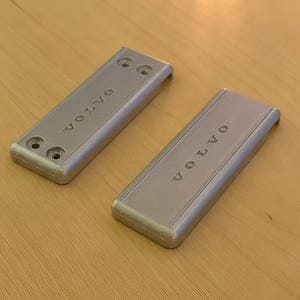 Peut inclure: Deux plaques rectangulaires en métal de couleur argentée avec le mot VOLVO en relief sur la surface. Une plaque présente deux trous de vis circulaires à chaque extrémité, tandis que l'autre a une surface lisse avec la marque en relief.