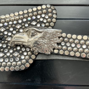 May include: A black leather belt featuring a large silver-toned eagle head buckle. The belt strap is heavily embellished with rows of round silver metal studs and clear, faceted rhinestones arranged in a dense, textured pattern.