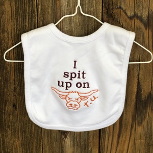 May include: White baby bib with brown embroidery that reads "I spit up on" with a cartoon longhorn steer head and the letters "X.U."