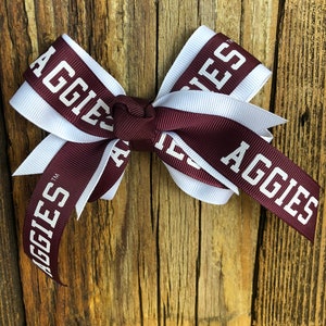 May include: A maroon and white bow with the word "Aggies" printed on it. The bow is tied with a knot in the center.