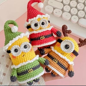 May include: Three crocheted figures resembling yellow characters with black goggles. One wears a red Santa hat and suit, one wears a green elf hat and outfit, and one wears brown reindeer antlers. Each has a white trim and a black belt with a gold buckle.