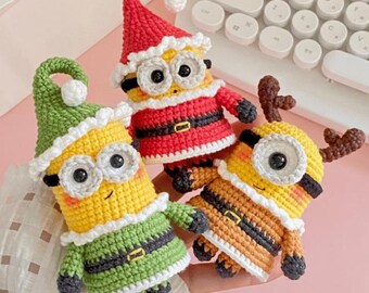 May include: Three crocheted figures resembling yellow characters with black goggles. One wears a red Santa hat and suit, one wears a green elf hat and outfit, and one wears brown reindeer antlers. Each has a white trim and a black belt with a gold buckle.