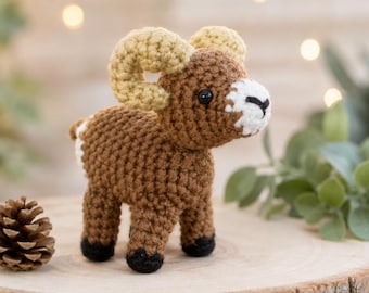 May include: A small crocheted ram figurine featuring a brown body, tan curled horns, a white muzzle with a black stitched nose, and black hooves. The stuffed animal is crafted with visible yarn stitches and has a rounded, compact shape.