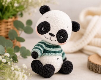 May include: A crocheted panda toy featuring a white head and body with black ears, eye patches, arms, and feet. The panda wears a green and white striped sweater and has a small embroidered 'x' on its belly. It has large dark eyes and a stitched mouth.