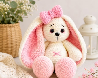 May include: A crocheted stuffed rabbit toy with cream-colored yarn for the body and head, featuring long ears with pink textured interiors. The rabbit has a pink crocheted bow on its head, black safety eyes, a black nose, and a pink stitched X on its belly.