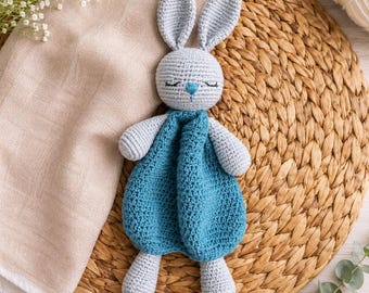 May include: A crochet security blanket featuring a light gray rabbit head with long ears, closed eyes, and a small blue nose. The body of the rabbit is a soft, textured teal blue blanket made of yarn, with light gray crochet arms and legs attached.