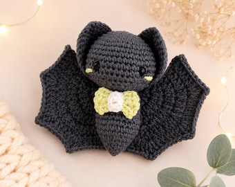 May include: A dark gray crocheted bat plush toy featuring large pointed ears, black safety eyes with small light yellow accents underneath, and a small bow tie at the neck made of light yellow and white yarn. The wings are wide and textured.