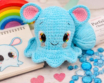 May include: A light blue crocheted plush toy with large black eyes featuring rainbow irises, pink inner ears, and a small embroidered smile. The plush has a rounded head and a ruffled base, crafted from soft yarn, sitting next to a drawing of itself.