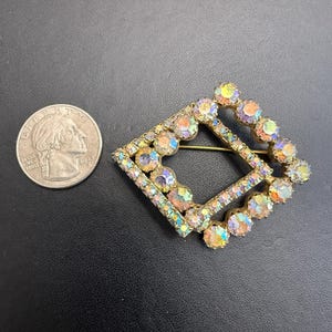 May include: A gold-tone metal brooch in a square frame shape, encrusted with iridescent, multi-colored rhinestones that reflect pink, blue, and yellow hues. The brooch is positioned next to a United States quarter coin for scale reference.