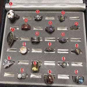 May include: A collection of eighteen assorted metal rings displayed in a gray velvet jewelry tray. The rings feature various gemstones, including white, purple, yellow, and blue stones, along with intricate metalwork, heart shapes, and oval settings.
