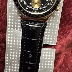 May include: A gold-tone chronograph wristwatch featuring a black dial with three gold sub-dials, a date window at the four o'clock position, and a black leather strap with a crocodile-embossed texture. The bezel includes a tachymeter scale.