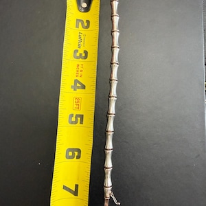 May include: A silver-toned metal bracelet with a segmented bamboo-like design, positioned next to a yellow measuring tape that shows the item is approximately 7.5 inches in length. The bracelet features a small clasp mechanism at one end.