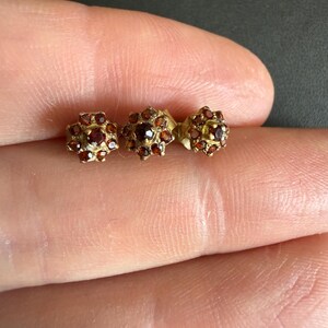 May include: Three small stud earrings featuring a cluster design. Each earring is crafted from a gold-toned metal and set with multiple round, deep red garnet gemstones arranged in a floral-like pattern with a central stone surrounded by six smaller ones.