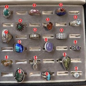 May include: A collection of eighteen assorted rings displayed in a gray velvet jewelry tray. The rings feature various designs, including oval gemstones, intricate metal bands, clear crystals, and patterned settings, each labeled with a red number.