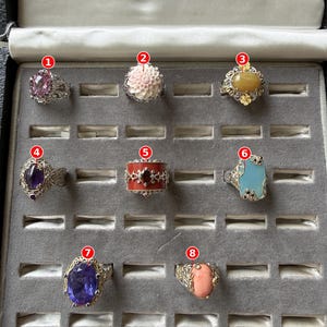May include: A collection of eight rings displayed in a gray velvet jewelry box. The rings feature various designs including a faceted pink stone, a carved floral motif, a yellow oval stone, a purple oval stone, a red band, a blue stone, a large purple stone, and a peach oval stone.