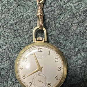 May include: A gold-tone pocket watch with a round face, featuring black Arabic numerals, gold hands, and a sub-dial for seconds. The dial is marked Paul Raynard, 17 Jewels, and Incabloc. A gold-tone metal chain is attached to the top bow.