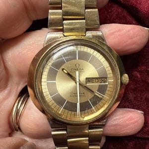 May include: A gold-tone Omega Genève Dynamic automatic wristwatch featuring a round case, a metallic gold and brown dial with a day-date window displaying WED 29, and a matching gold-tone link bracelet with a secure fold-over clasp mechanism.