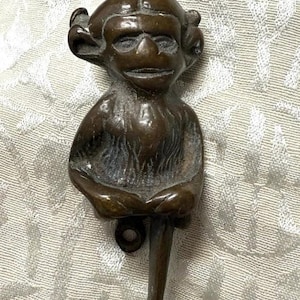 May include: A dark bronze-colored metal door knocker shaped like a seated creature with pointed ears, a textured chest, and a single extended leg. The figure features a loop at the top and a circular mounting hole at the bottom for attachment.